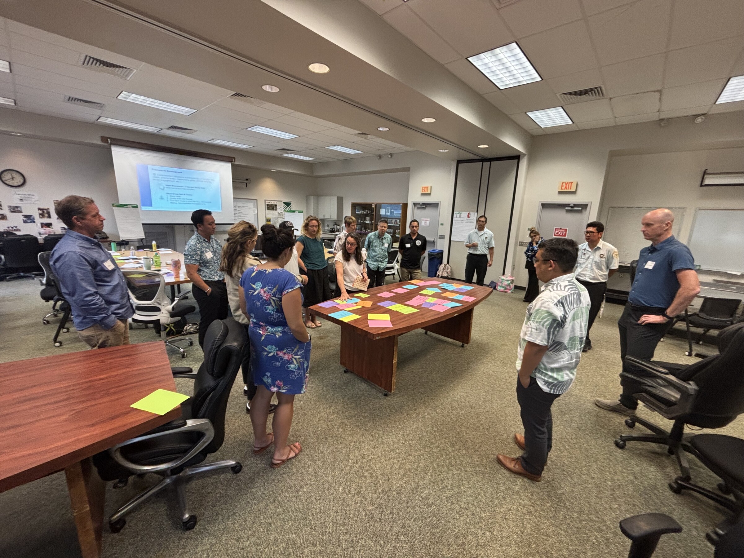 Several people standing in a room with colorful paper on a table and a projector screen in the back with a presentation.